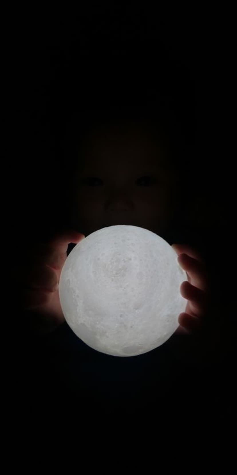 Child's hands hold a glowing moon orb in darkness with subtle eyes visible, providing soft light in a black scene.