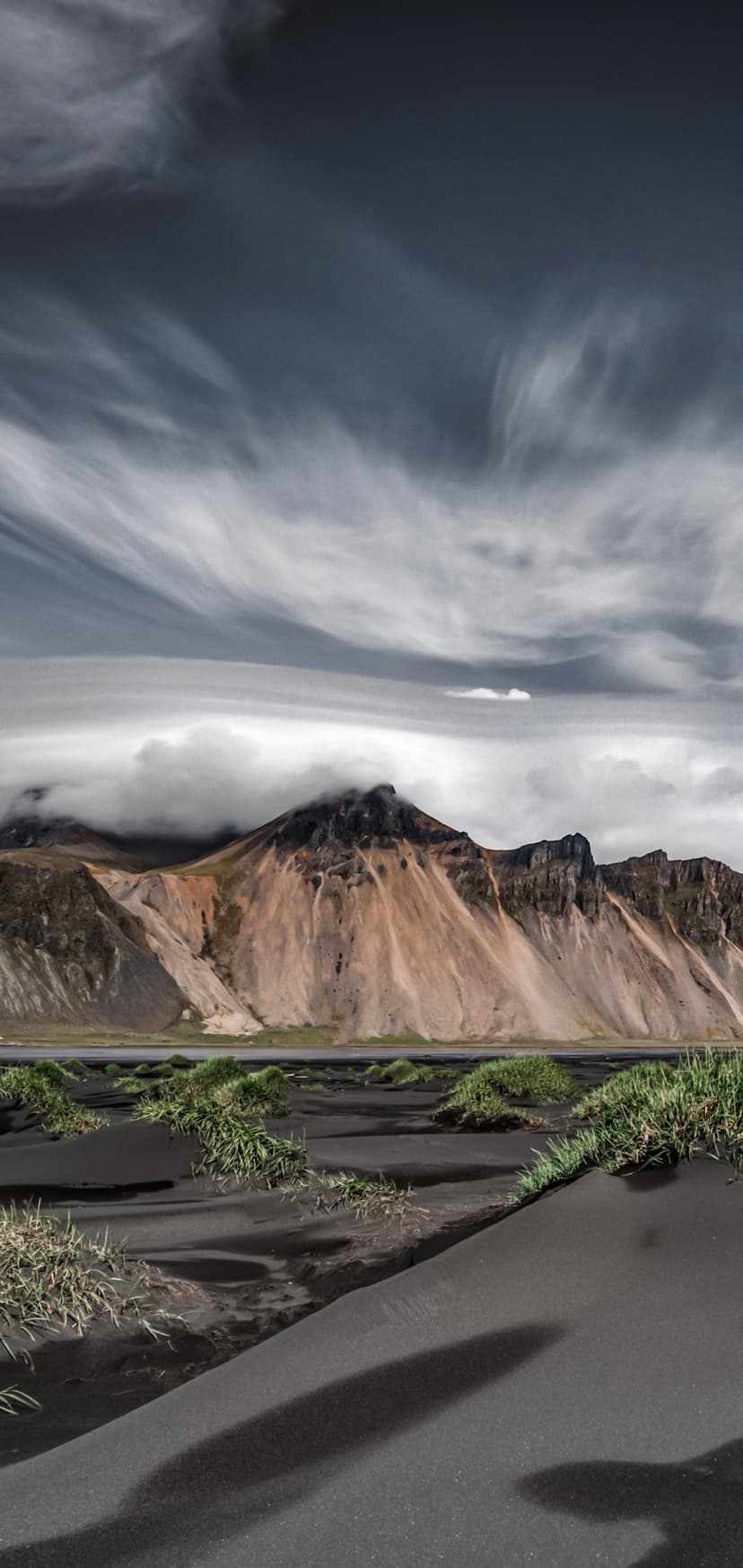 Vestrahorn mountain peaks rise behind black sand dunes and green grass tufts under a swirling, cloudy sky.