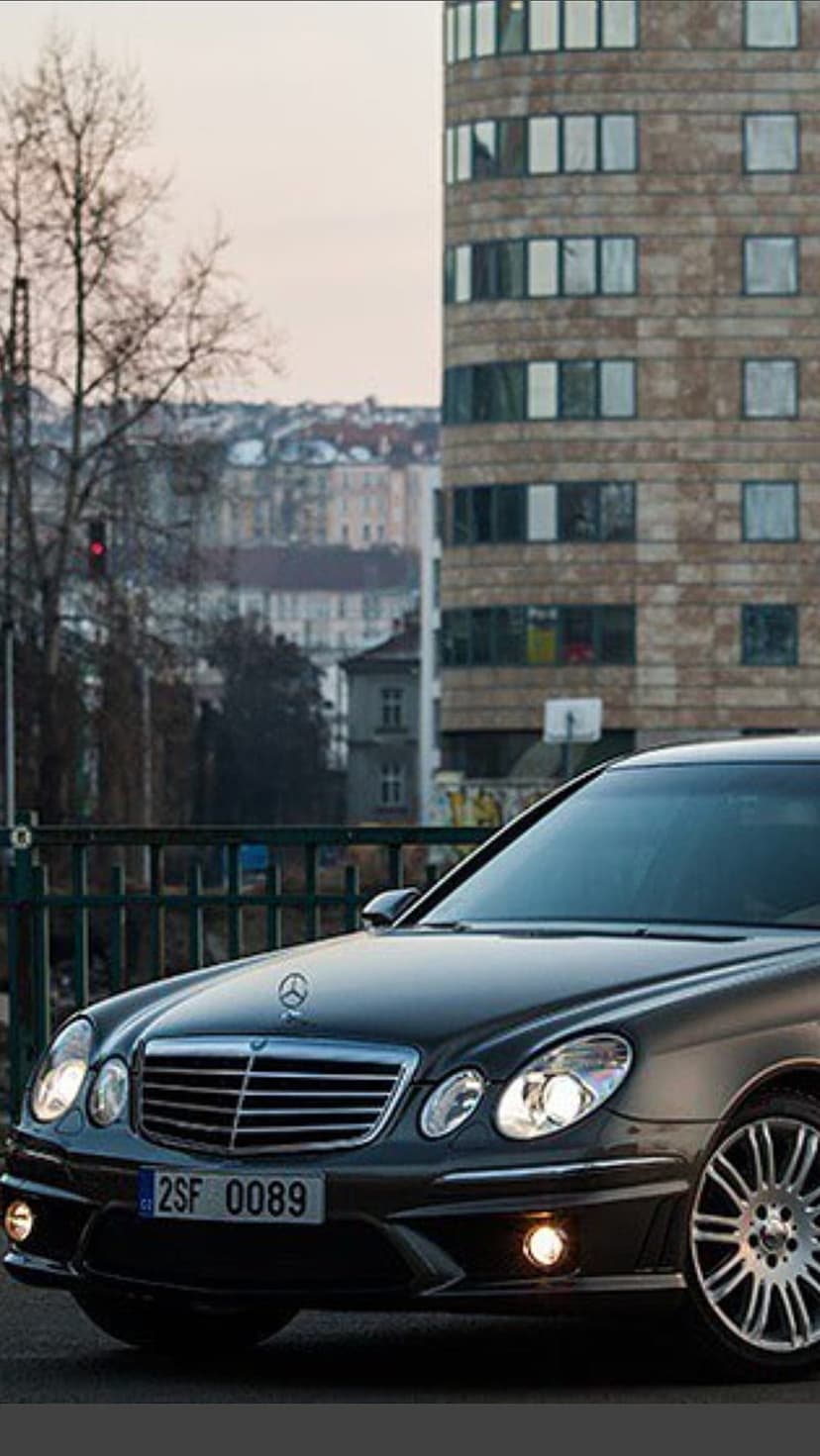 Mercedes E-Class sedan parked on a city street at night with glowing headlights and a stone building background.