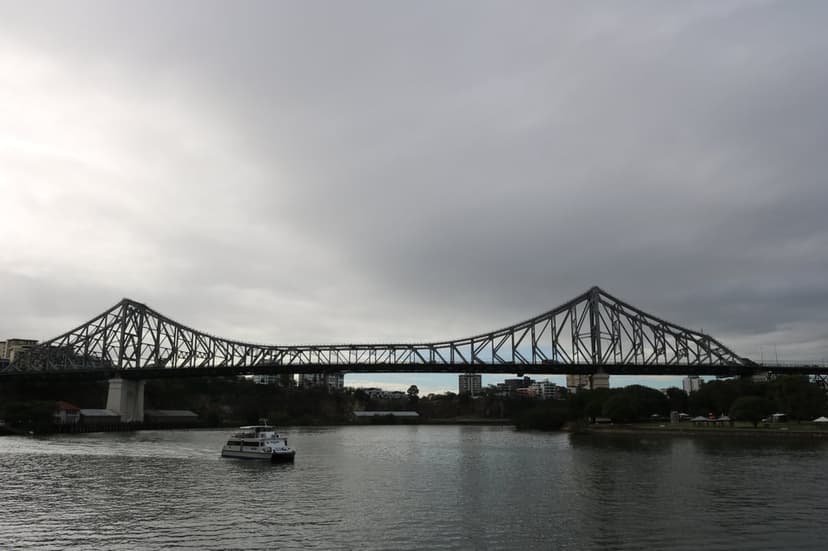 Story Bridge spanning the Brisbane River with a ferry on the water under a dramatic grey cloudy sky.