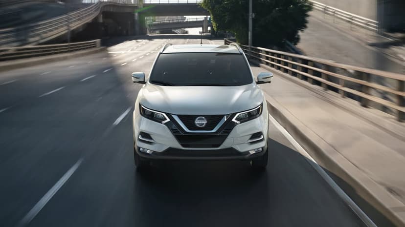 Front view of a white Nissan crossover SUV driving on a sunlit city highway bridge with urban structures.