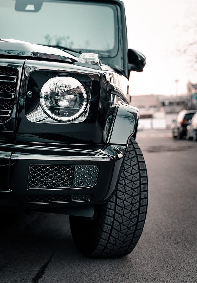 Circular LED headlight of a black G-Wagon beside a textured tire tread with sharp automotive reflections.