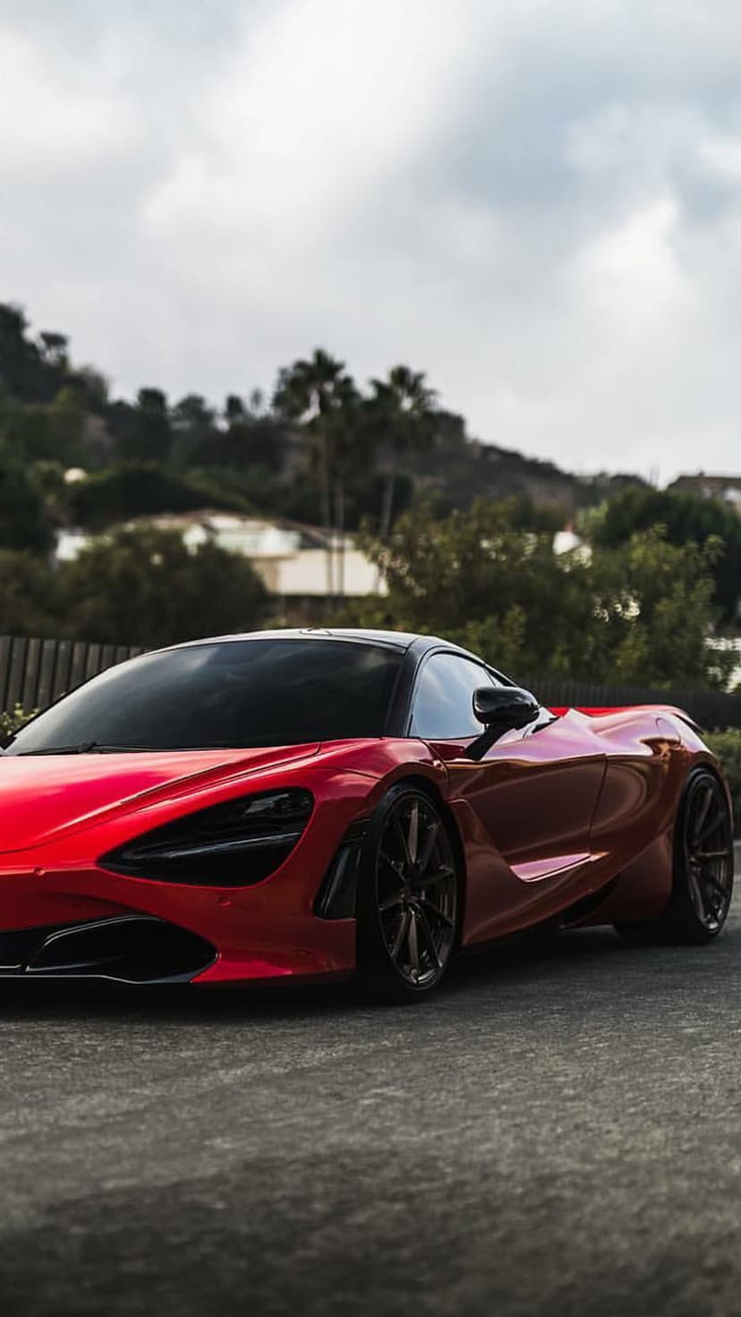 Red McLaren 720S supercar parked on a road with lush green hills and a cloudy sky in the background.