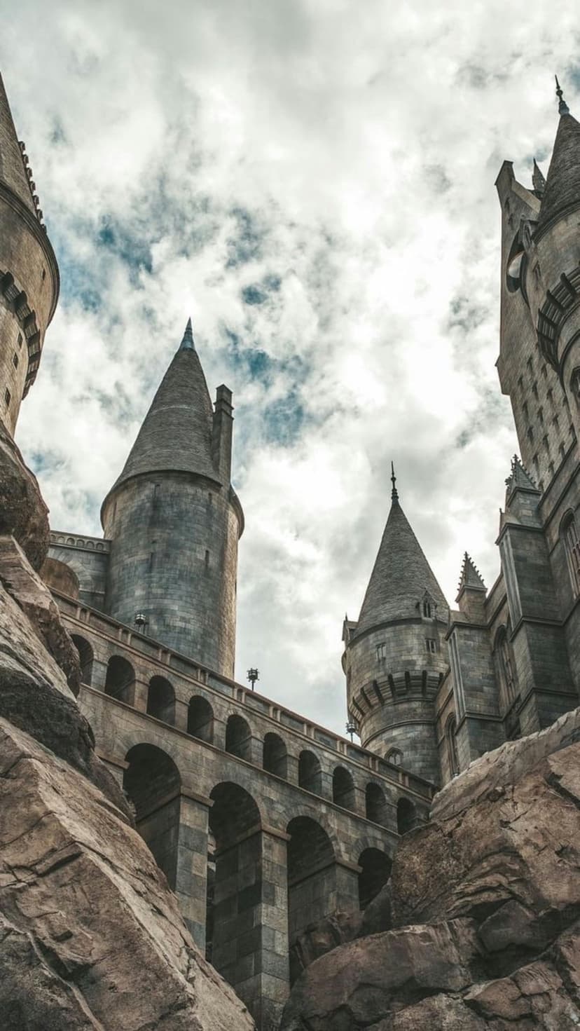 Hogwarts Castle spires and arched stone windows rise from rugged rocky cliffs against a dark and moody cloudy sky.