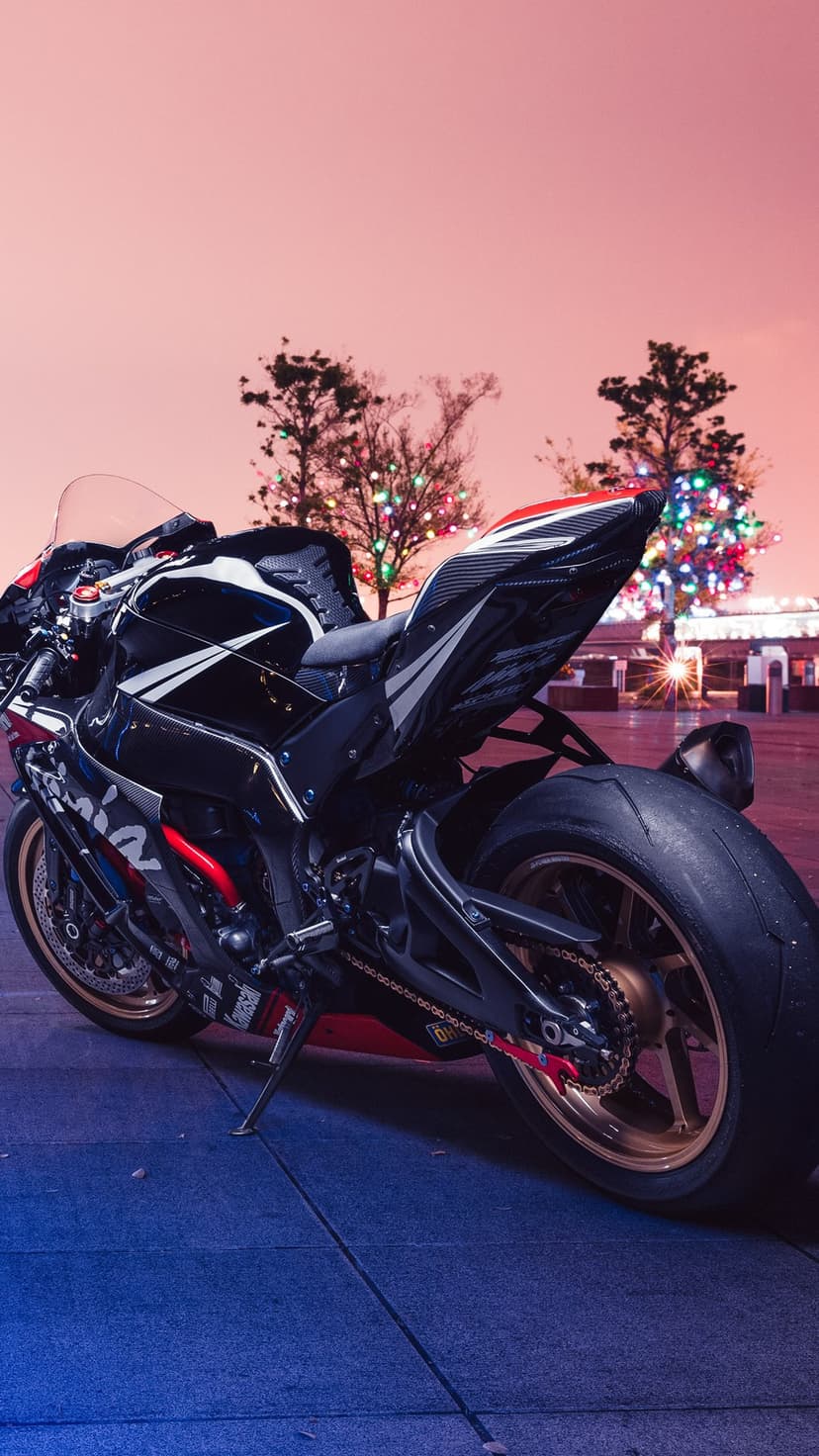 Black sportbike with white racing stripes and a gold wheel parked near trees glowing with colorful holiday lights.