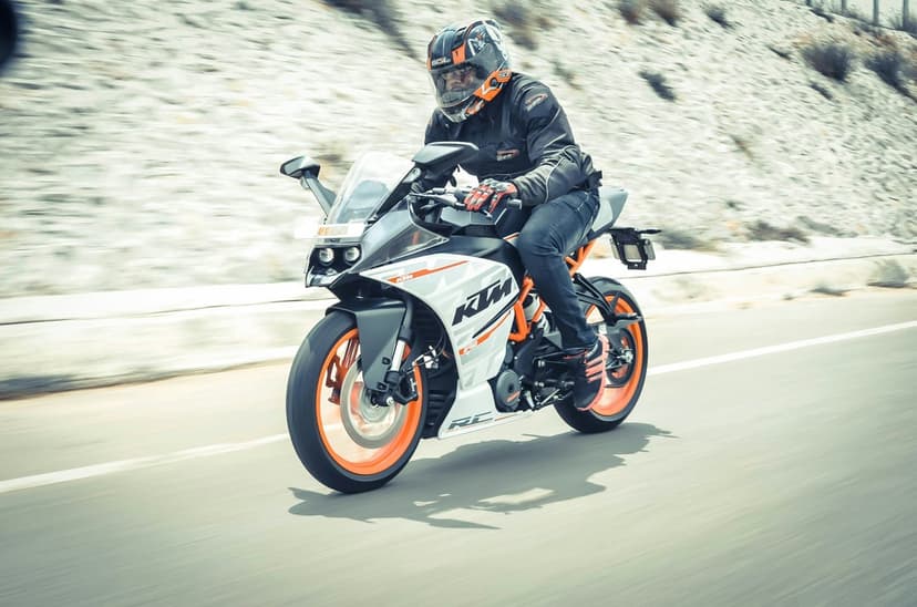 Motorcyclist in a full helmet leans on an orange KTM RC sportbike while riding along a blurred hillside road.