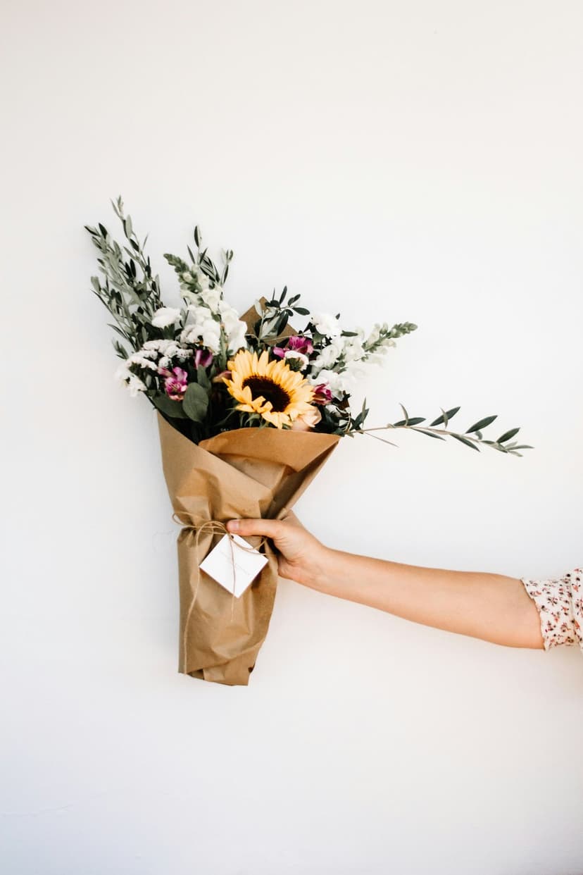 Person's hand gripping a sunflower bouquet wrapped in brown paper with a small tag against a white backdrop.
