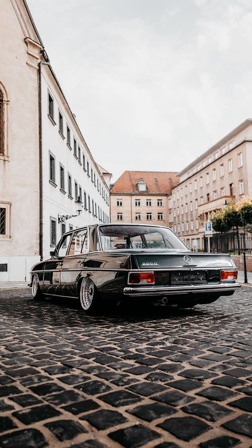 Dark Mercedes sedan with chrome rims parked on a cobblestone road in front of historic white European buildings.