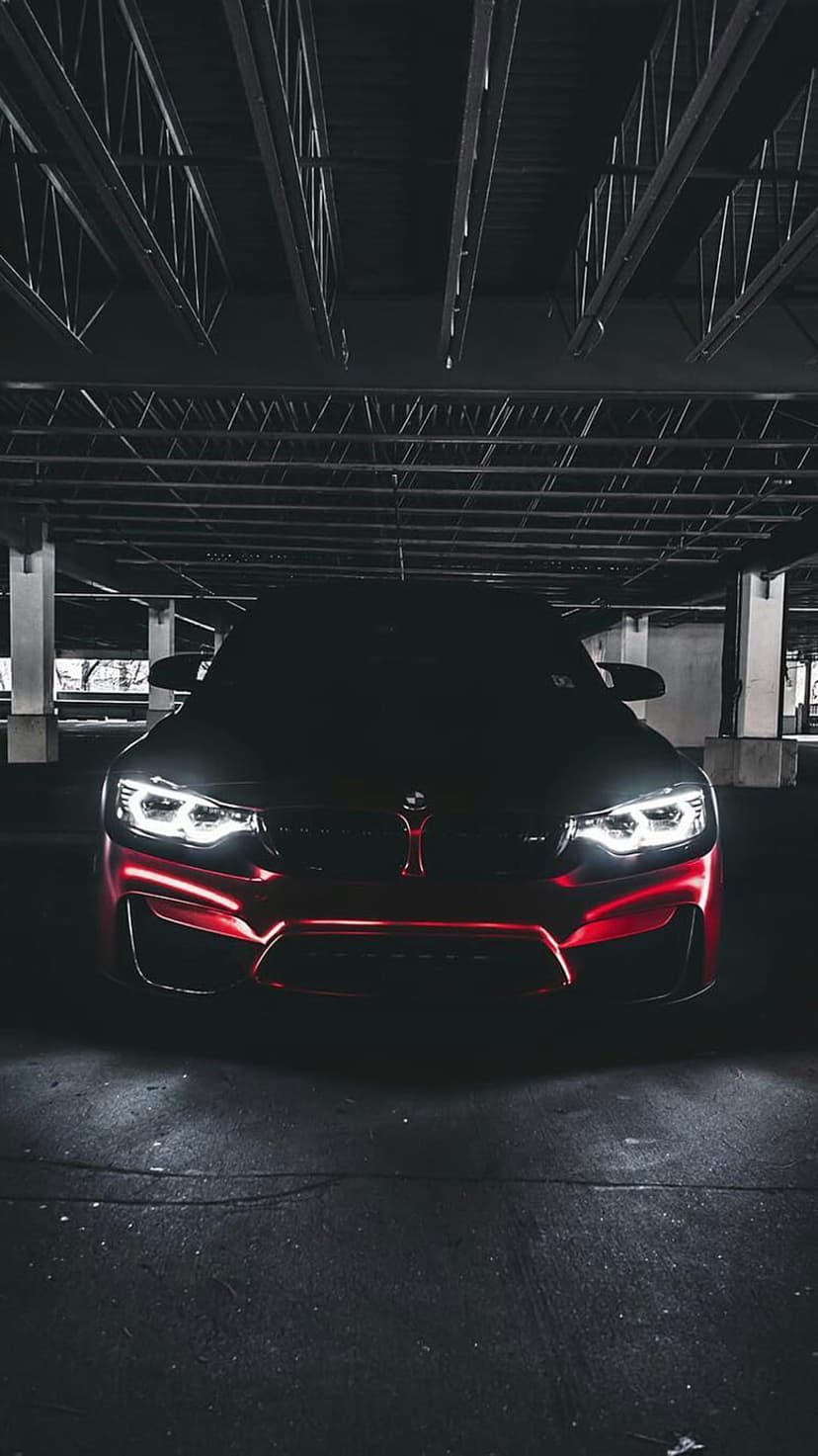 BMW M4 sports coupe with glowing red headlights parked in a dark industrial garage with dramatic red lighting.