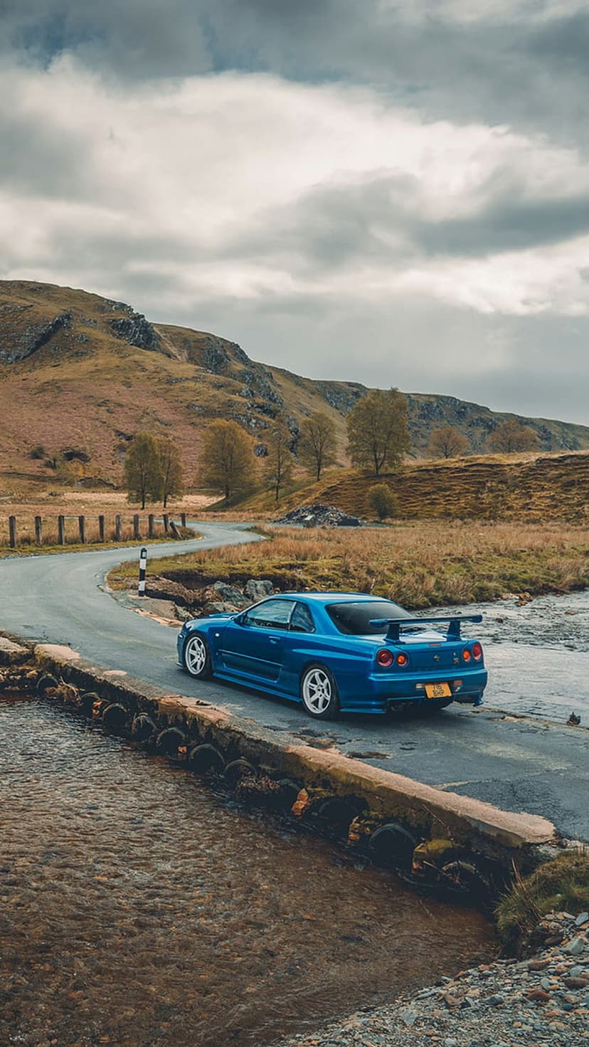 Nissan Skyline GT-R R34 in blue crosses a small bridge among autumn hills and a dramatic, overcast sky.
