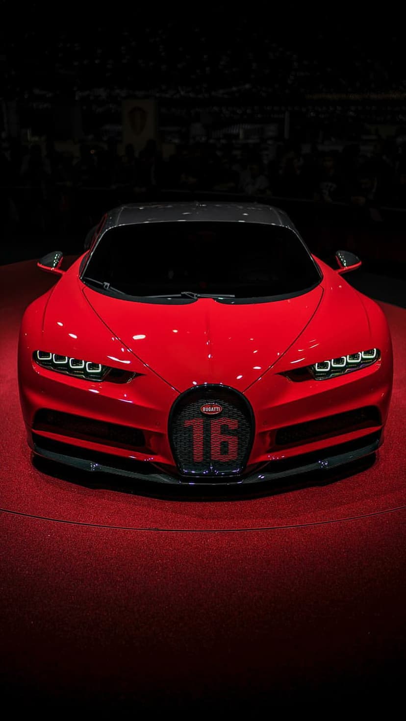 Red Bugatti Chiron hypercar on a red carpet with glowing headlights and 16 emblem on the horseshoe grille.