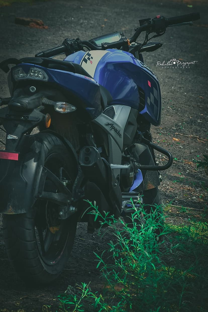 Blue Apache sportbike with checkered tank accents sits on a dirt path surrounded by blurred green foliage.