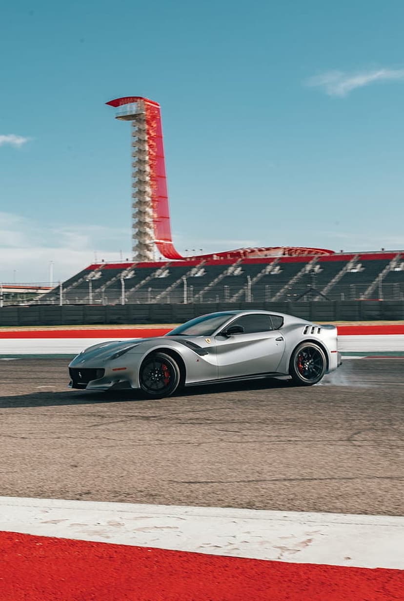 Silver Ferrari F12 TDF supercar driving fast on a race track with a red and white tower in the background.
