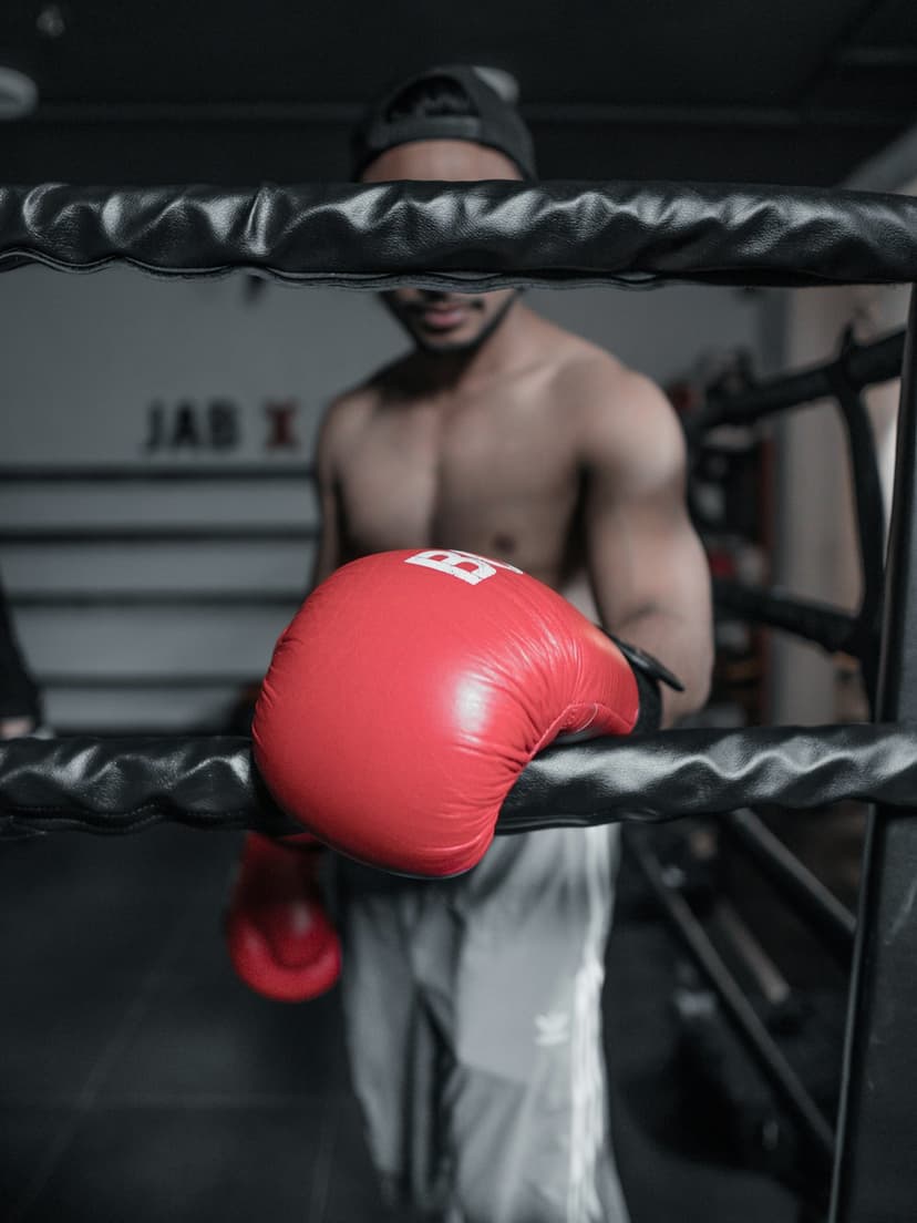 Shirtless male boxer wearing red gloves and a black cap rests against the padded ropes of a boxing ring gym.