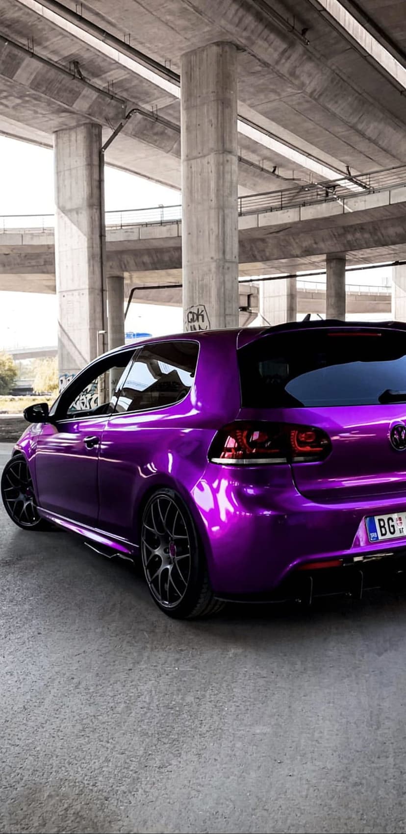 Purple Volkswagen Golf R with aftermarket wheels parked under a grey concrete overpass at a rear angle.