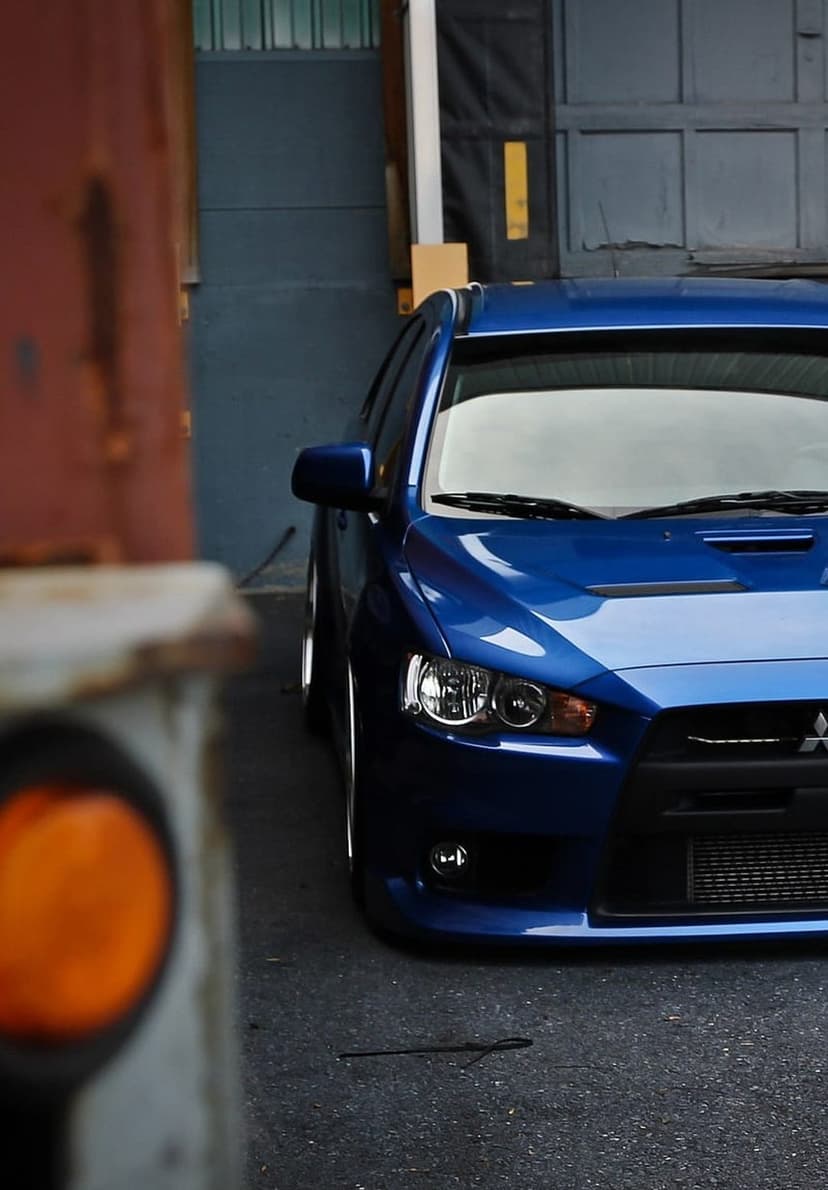 Blue Mitsubishi Lancer Evolution with a hood scoop and sharp headlights parked in an industrial urban setting.