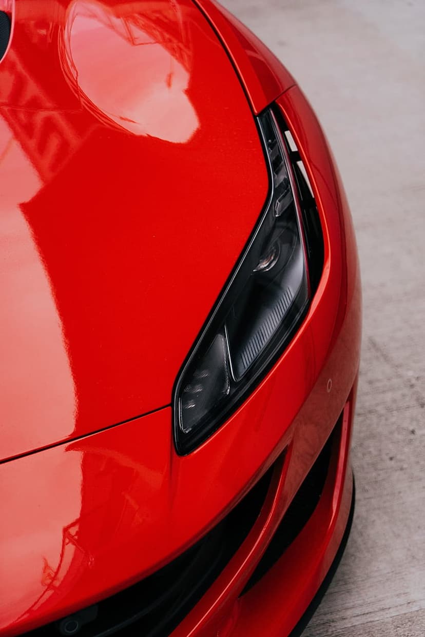 Red Ferrari headlight and hood showing reflections of light on polished Rosso Corsa paint and glass housing.