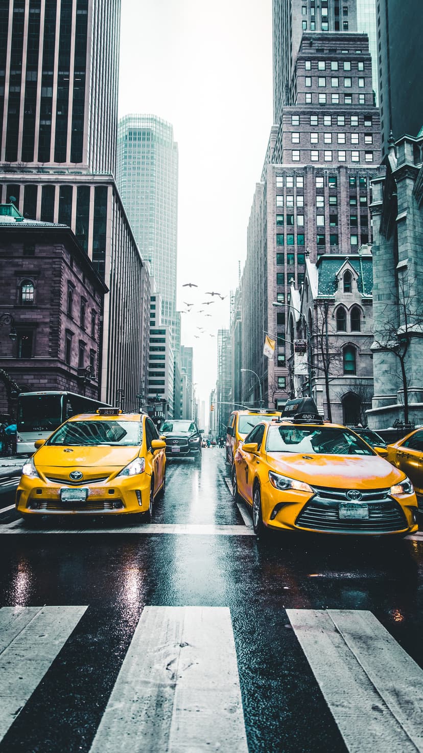 Yellow cabs drive across a rain-slicked city intersection surrounded by towering buildings and an overcast sky.