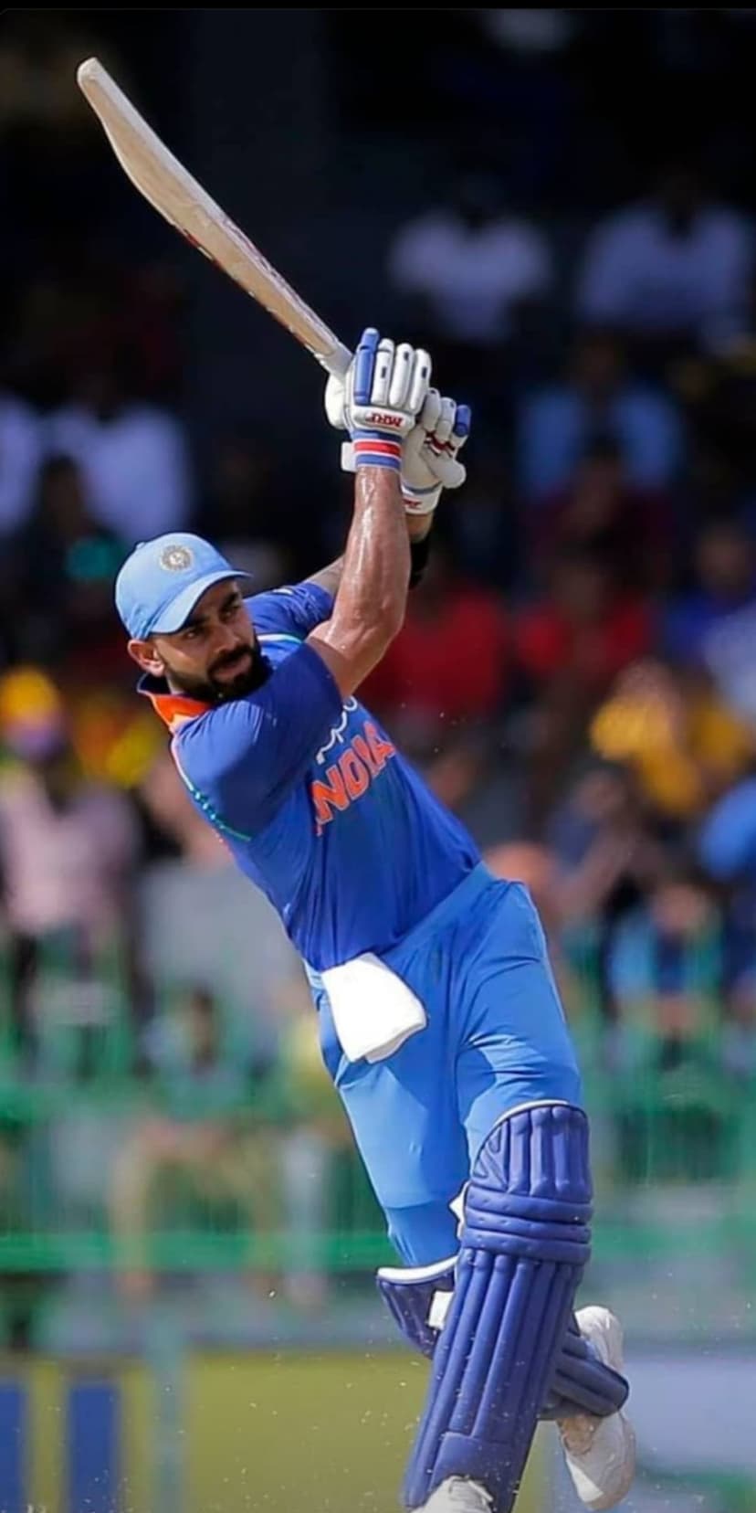 Virat Kohli in a blue jersey holds a cricket bat high while focusing on a ball during an outdoor cricket match.