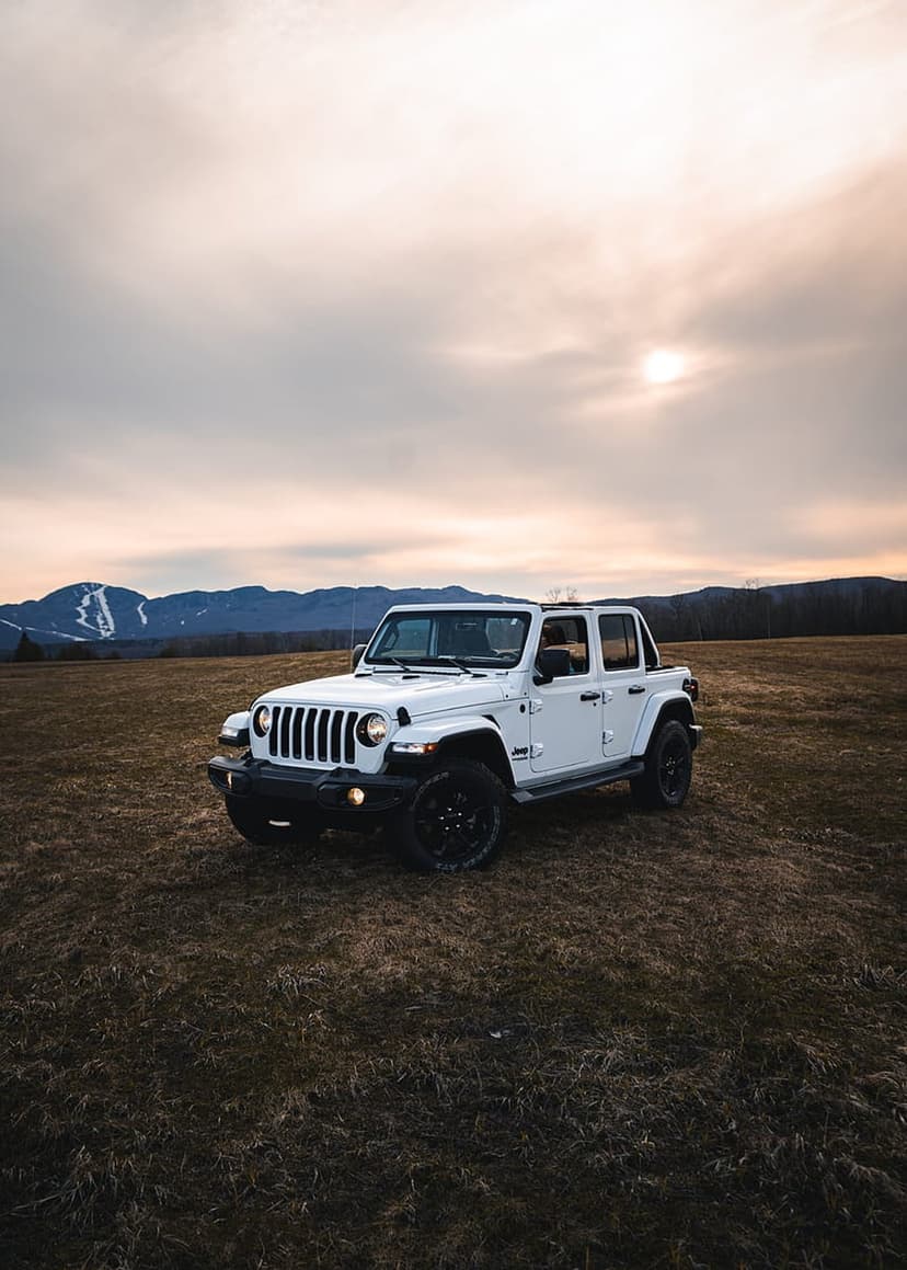 White Jeep Wrangler sits in a grassy alpine field before sharp mountain ridges under a soft sun-drenched sky.