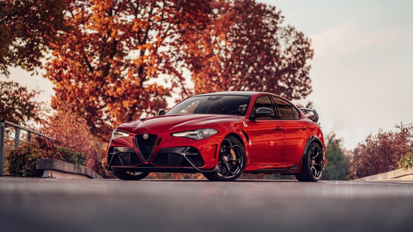 Red Alfa Romeo sports sedan on asphalt with orange autumn trees and soft golden lighting in the background.