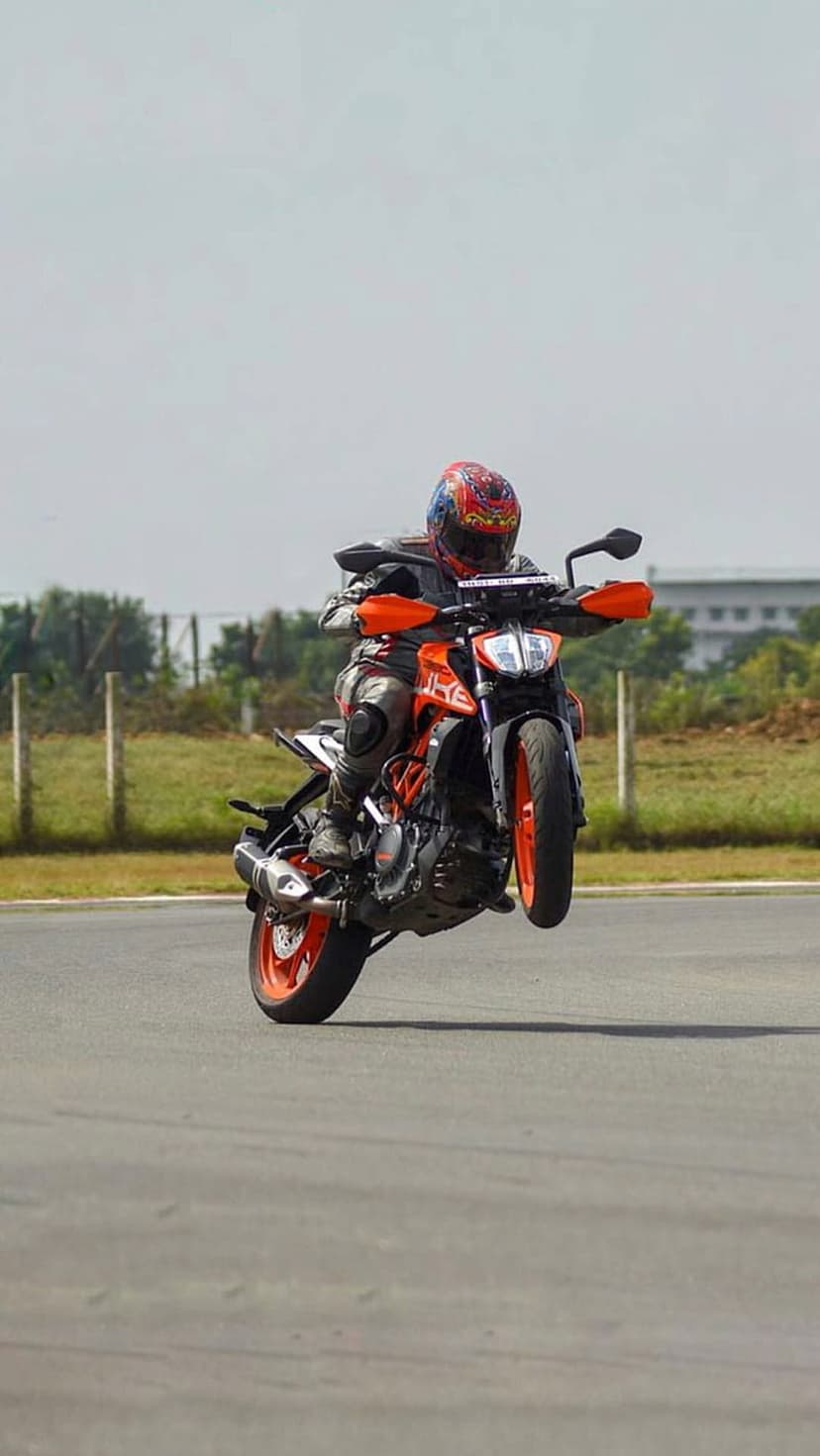 Professional motorcyclist performs a vertical wheelie on an orange KTM sportbike along a paved racing track.