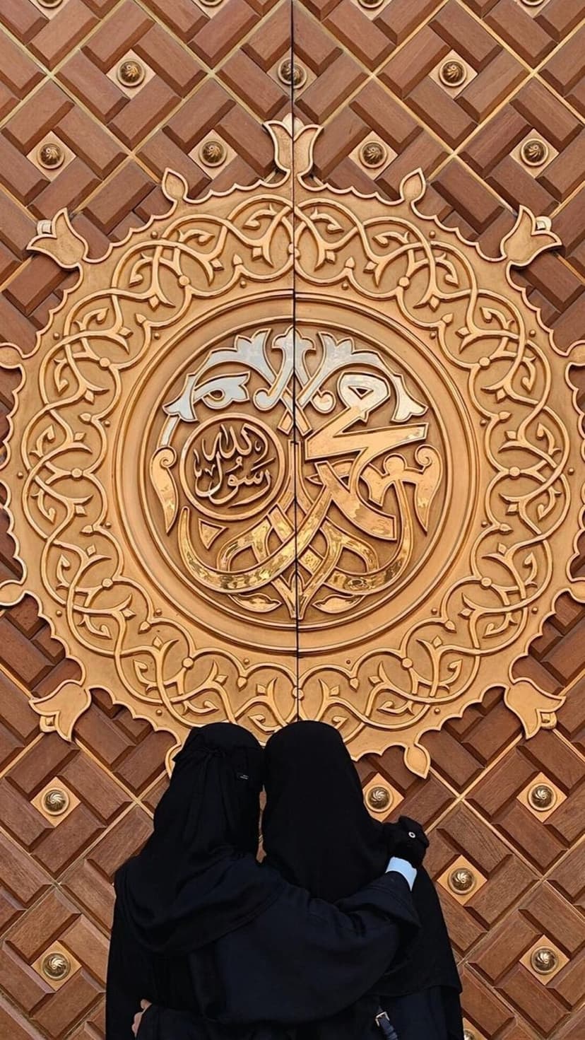 Two women in black abayas embrace before the ornate geometric wooden doors of the Prophet's Mosque in Medina.