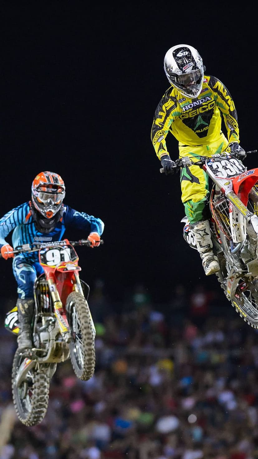 Motocross riders in blue and yellow gear jump their dirt bikes mid-air against a dark night stadium sky.