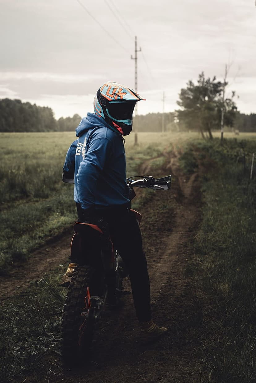 Motocross rider wearing a blue hoodie and helmet sits on a bike on a grassy dirt path under an overcast sky.