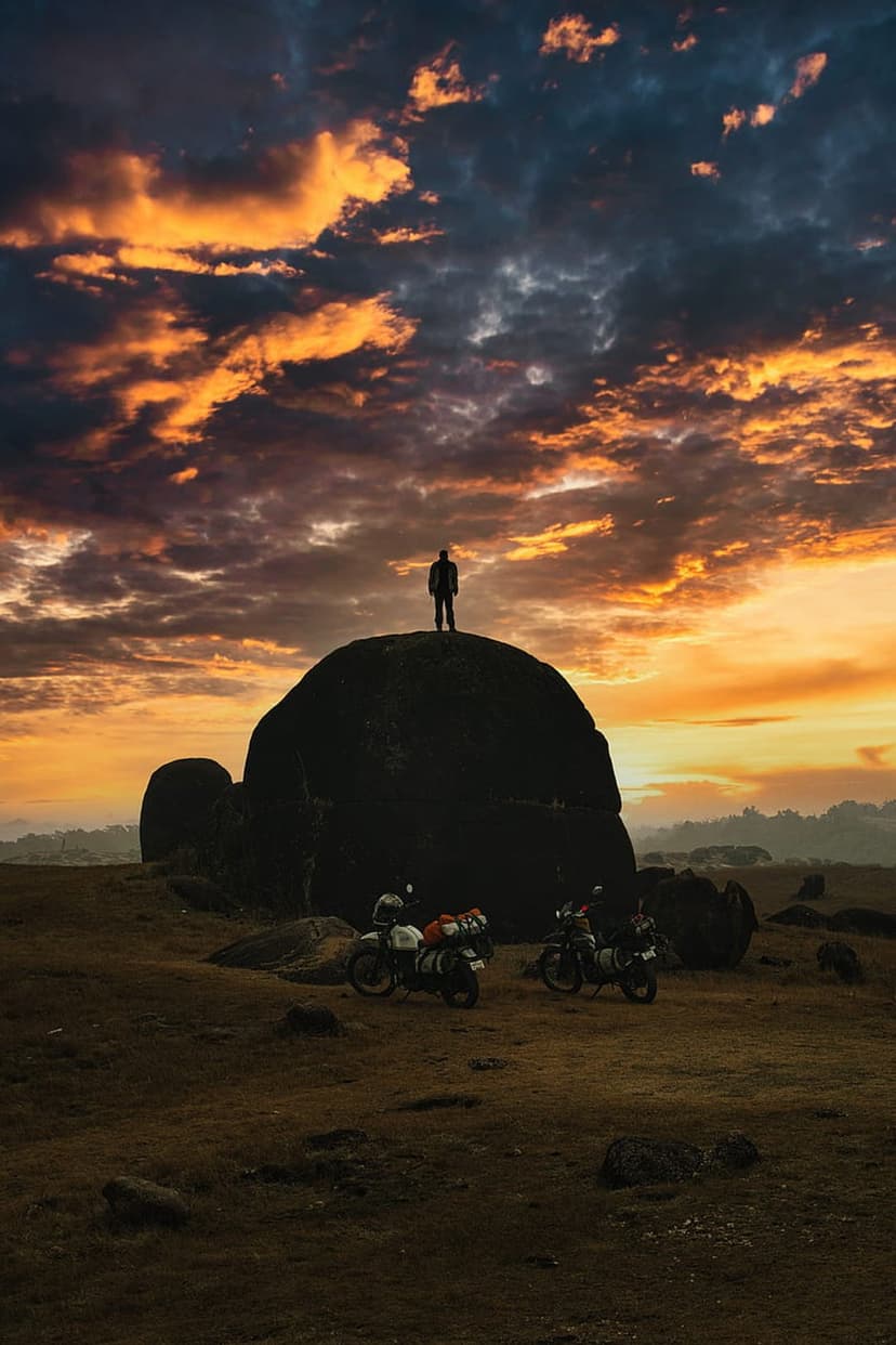 Man stands on a boulder silhouetted against an orange sunset sky near two parked touring motorcycles.