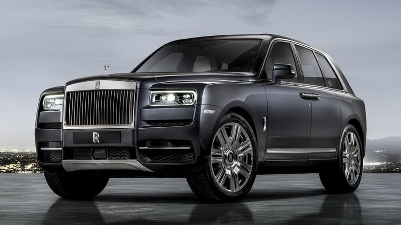 Dark grey Rolls-Royce Cullinan SUV with glowing headlights on a reflective floor against a night city.