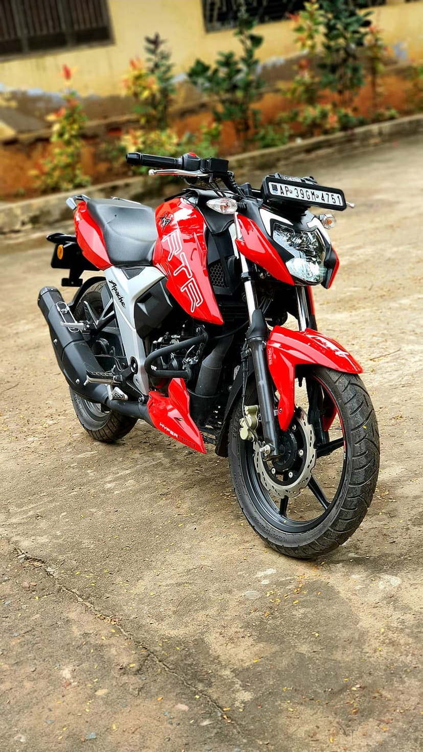 Red Apache RTR motorcycle parked on cracked concrete with a blurred background of green plants and a building.