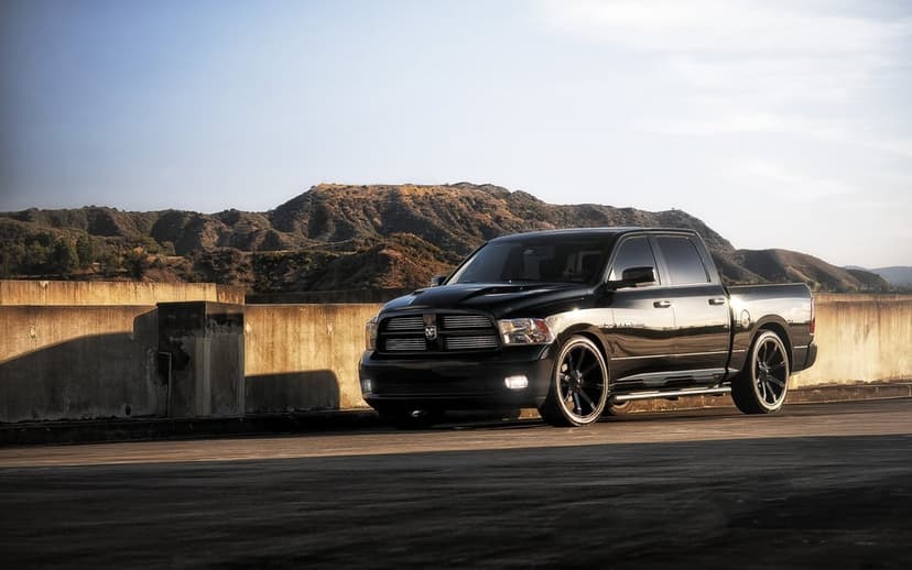 Black custom Dodge Ram truck with large rims parked on a concrete ledge overlooking brown hills at sunset.
