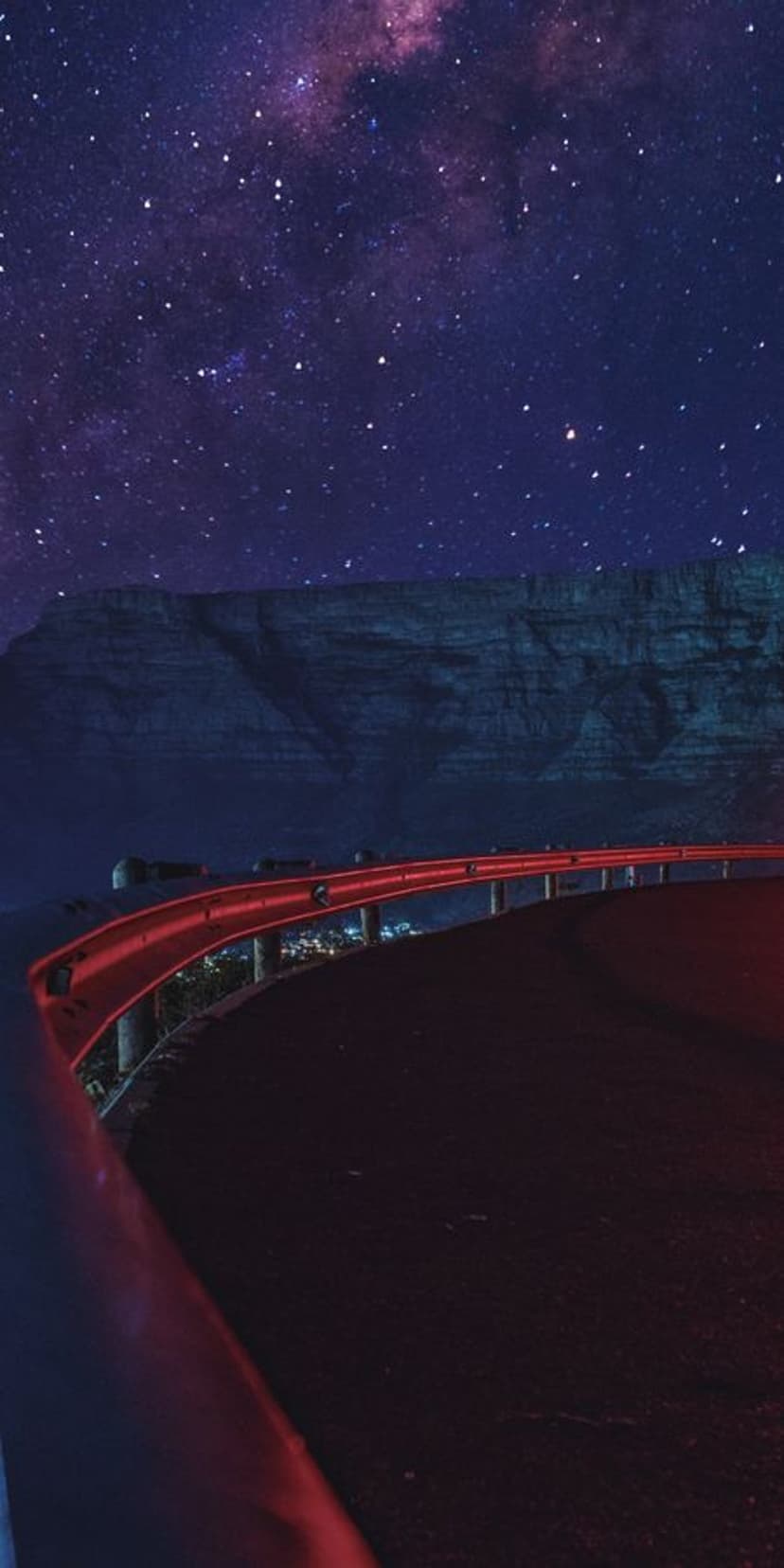 Red guardrail curves along a dark asphalt road under a purple Milky Way galaxy filled with glowing star clusters.