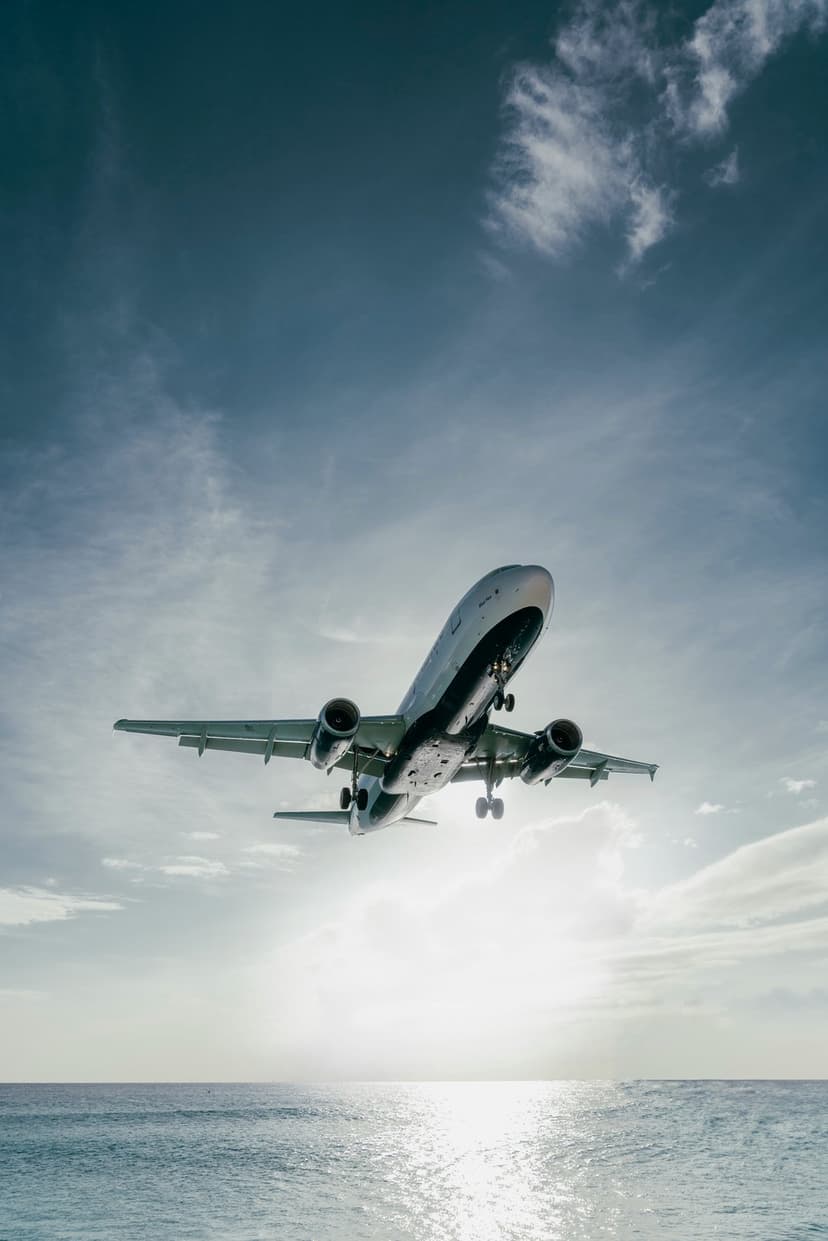 Commercial airplane climbing through a bright sunny sky over a blue ocean with shimmering water reflections.
