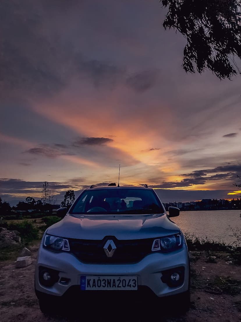 Renault Kwid hatchback parked at a lakeside during a purple sunset with colorful clouds reflecting on water.
