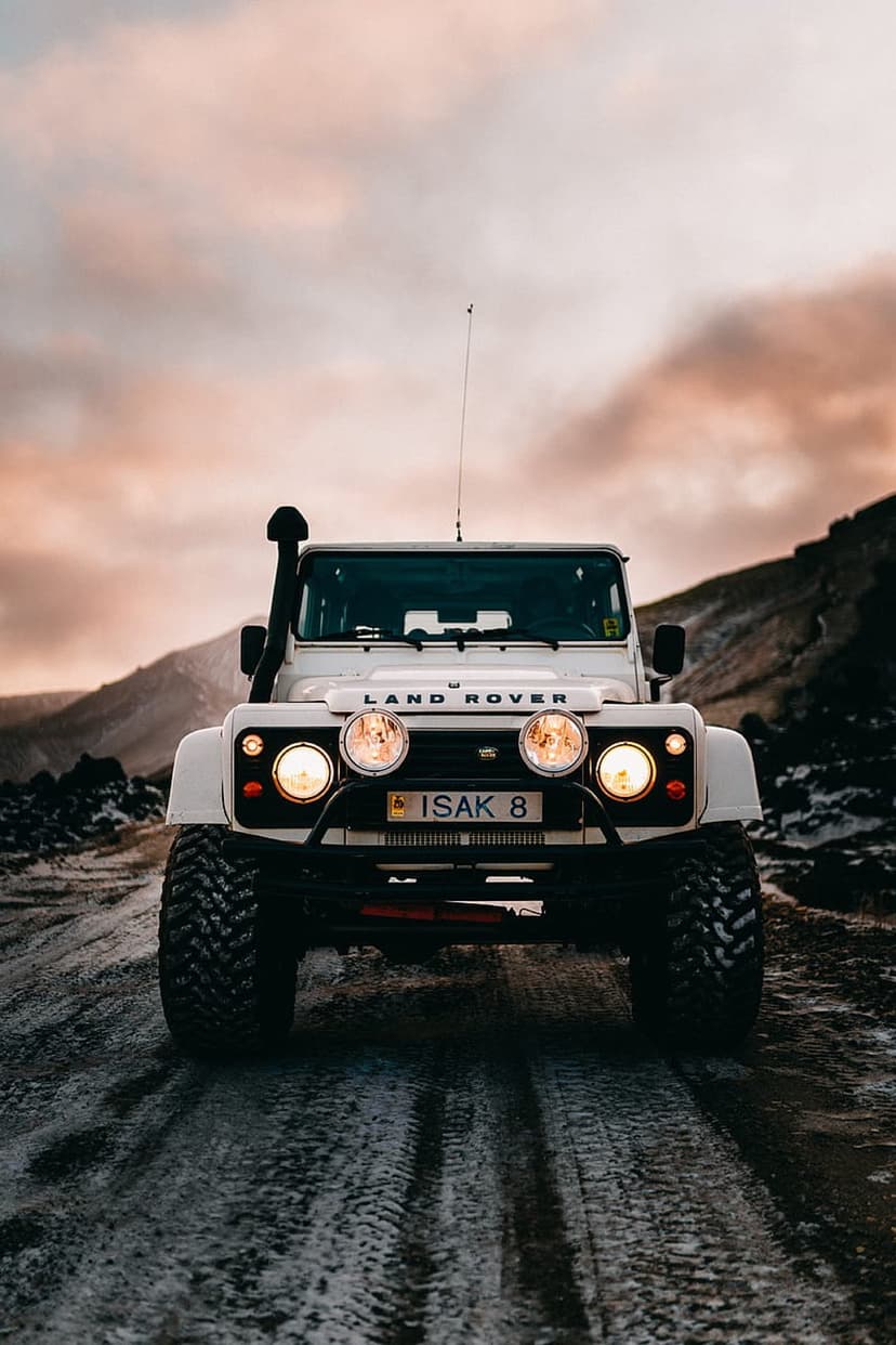 White Land Rover Defender 4x4 with snorkel driving on a wet, icy road toward a colorful mountain sunset.