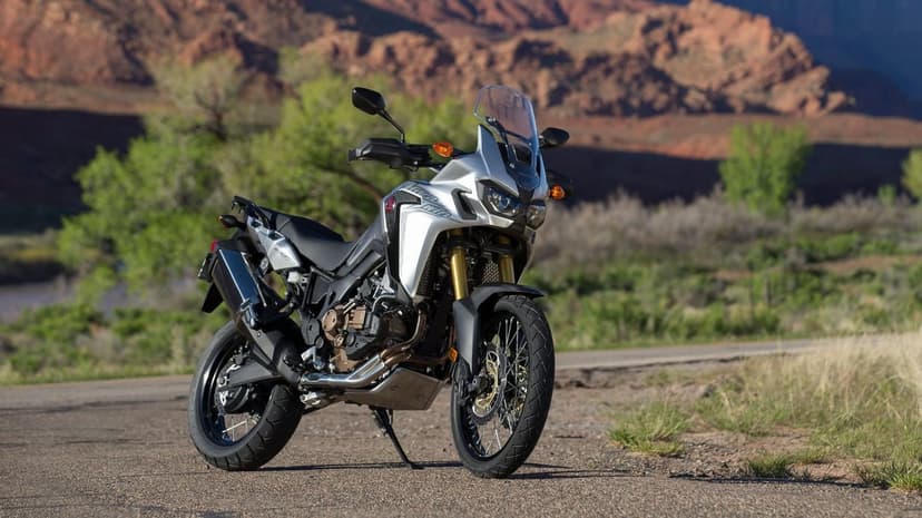 Silver Honda Africa Twin motorcycle parked on an asphalt road with red rock canyons and green trees behind.