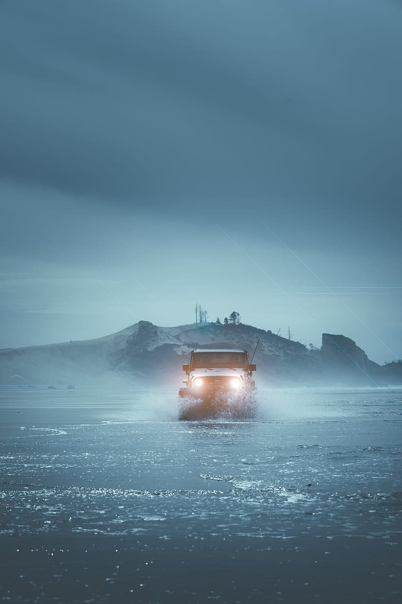 Jeep Wrangler with glowing headlights drives through splashing ocean waves on a misty, desolate coastal beach.