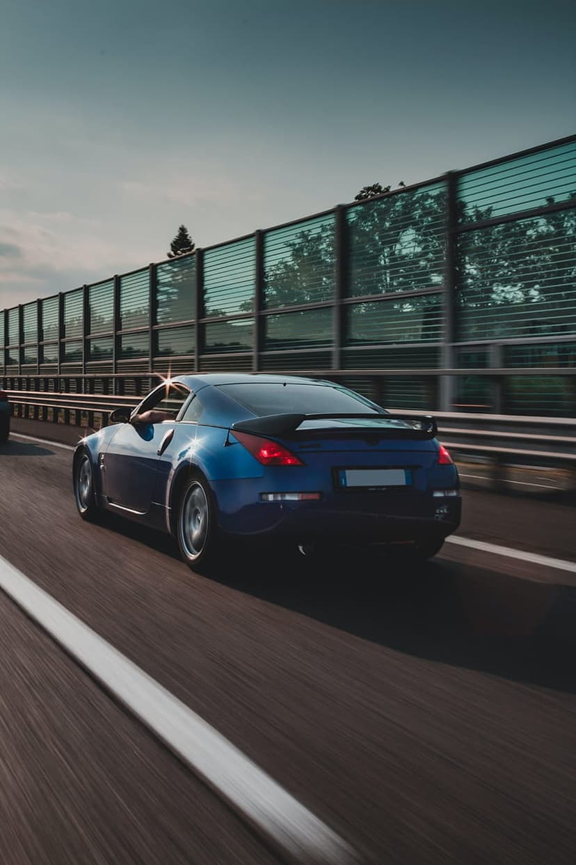 Blue sports car driving fast on a highway with motion blur, reflections of trees, and bright summer sunlight.
