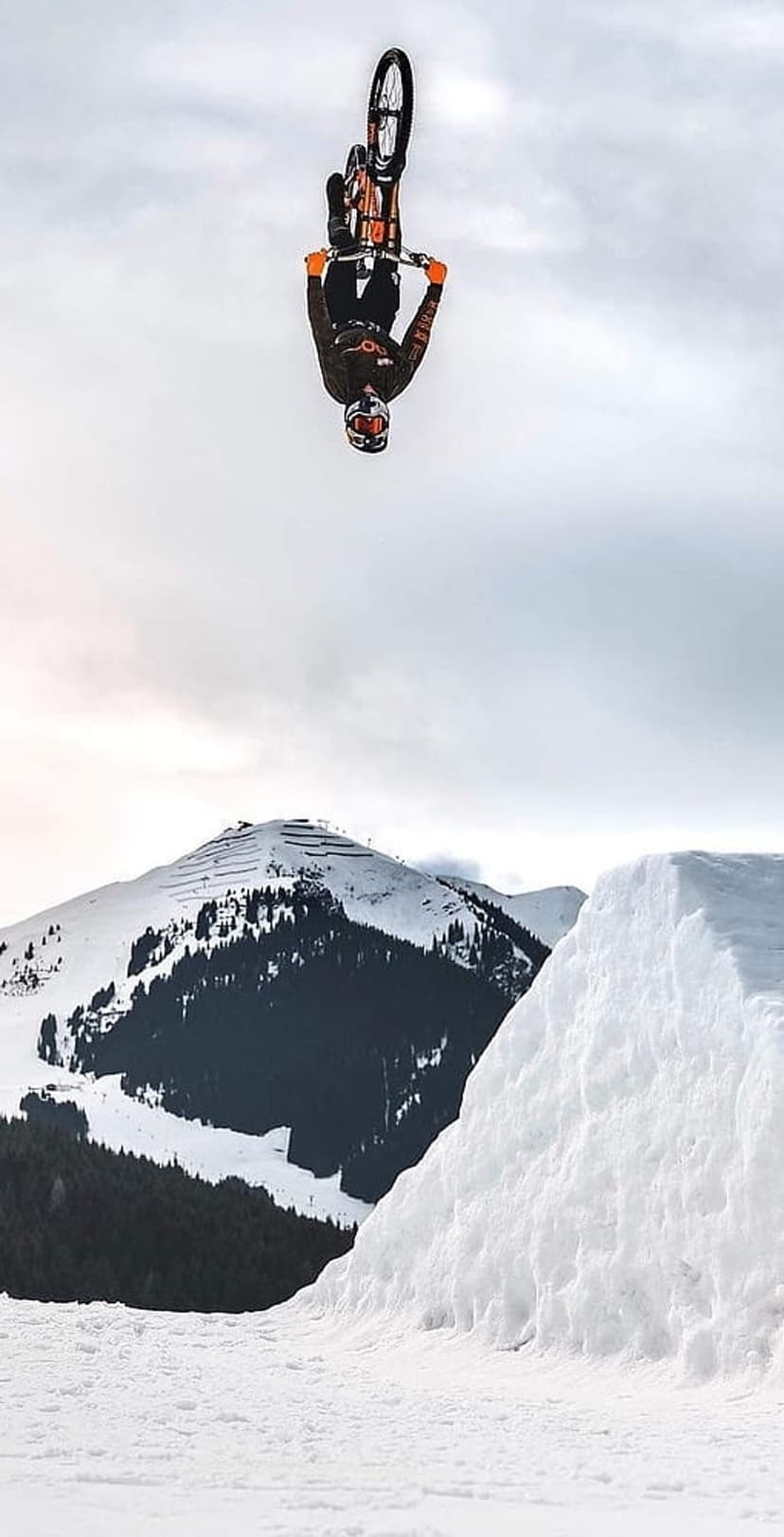 Mountain biker performs a backflip over a snowy ramp against a cloudy sky and pine trees in a winter landscape.