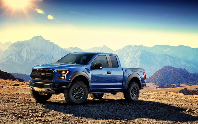 Blue Ford Raptor pickup truck parked on rocky desert terrain with mountains and sun flare in the background.
