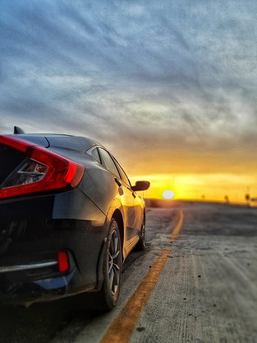 Black car parked on a paved road at sunset with orange light reflecting on its body under a cloudy evening sky.