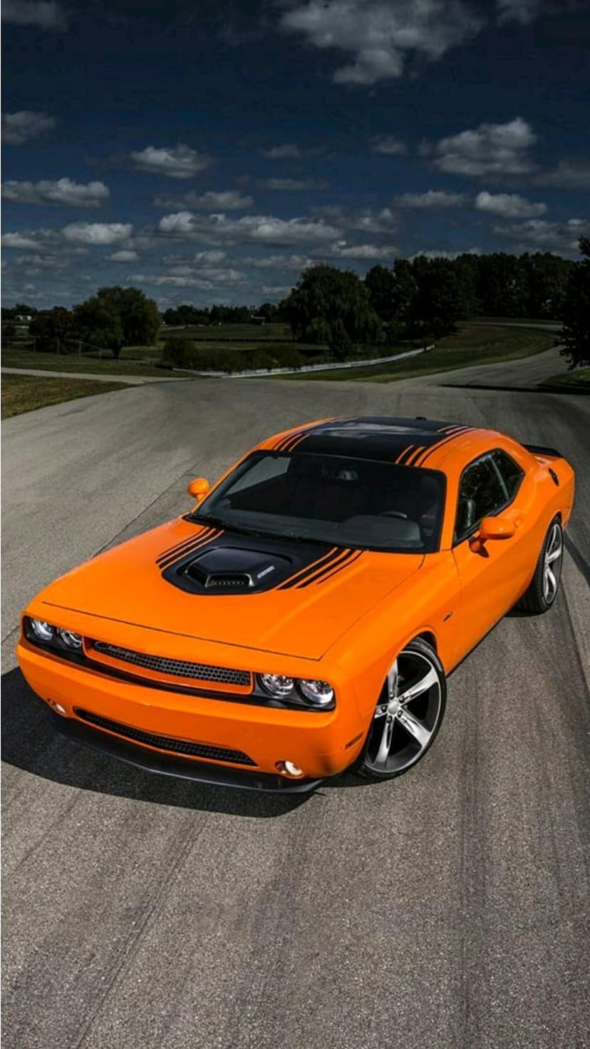 Orange Dodge Challenger R/T muscle car with black stripes on an asphalt road with trees and a cloudy sky.