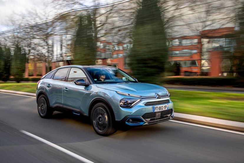 Light blue Citroën ë-C4 electric car driving on an asphalt road with motion blur and city trees in background.