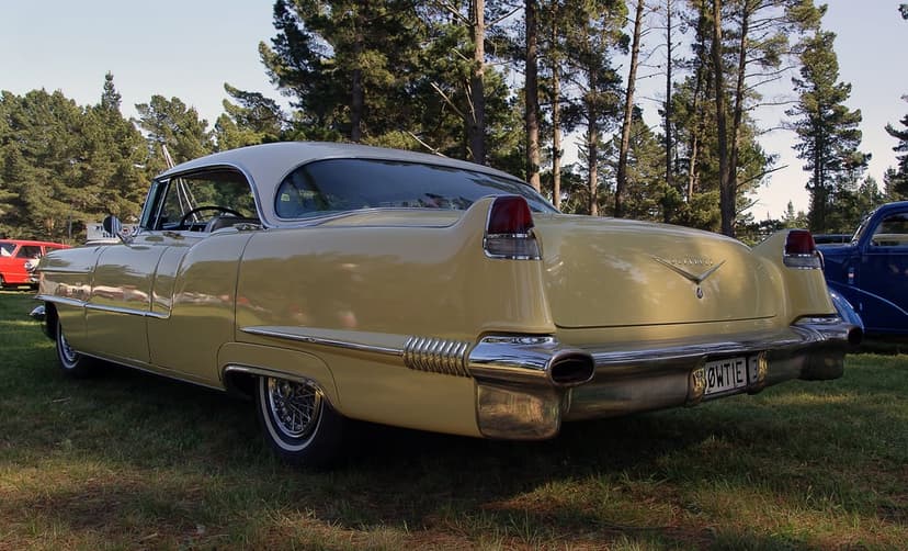 Yellow Cadillac Coupe Deville rear fins and chrome bumper parked on a grassy field with trees in the background.