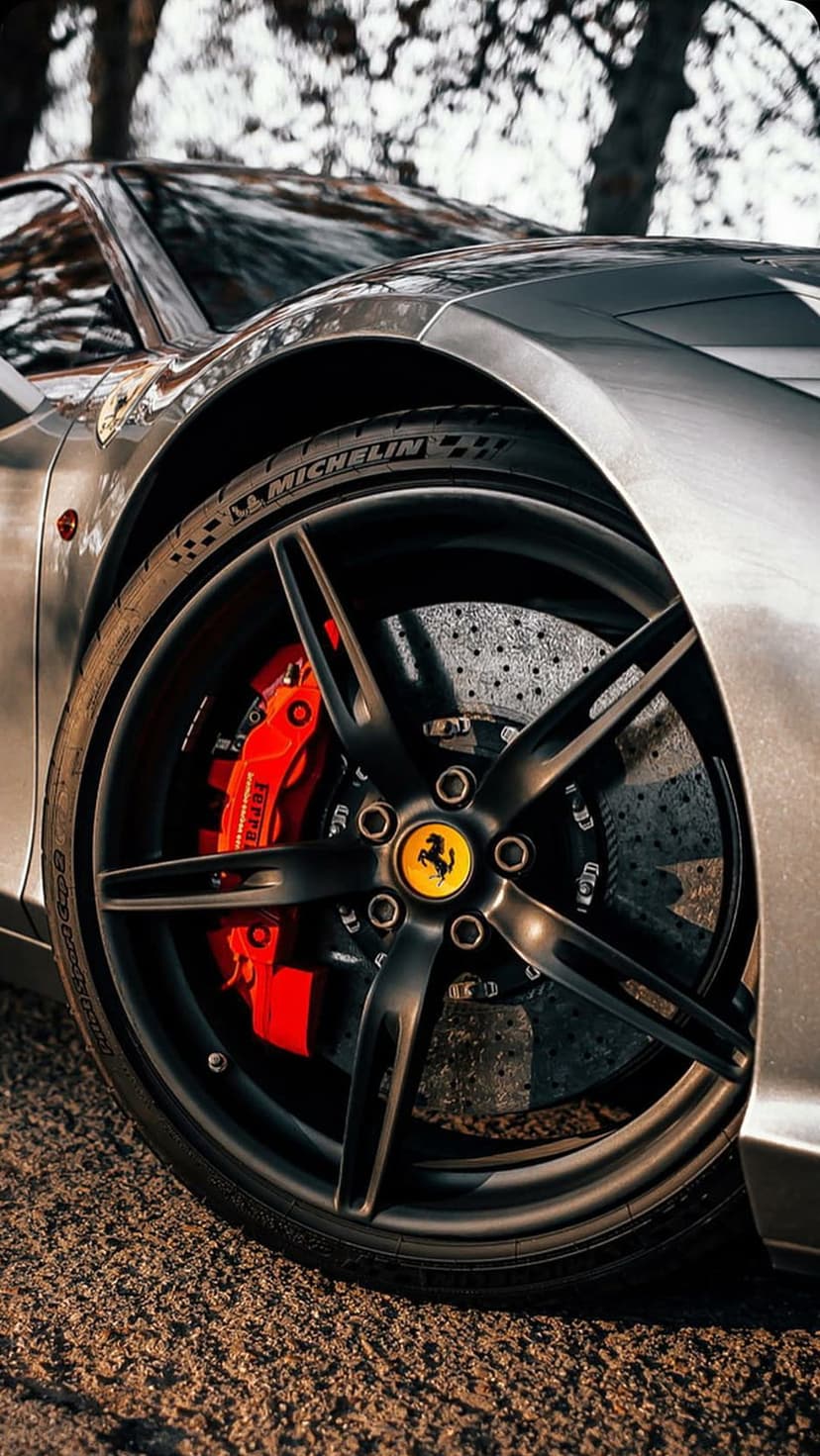 Ferrari black alloy wheel with red brake calipers, drilled metal discs, and Michelin tire in a low-angle shot.
