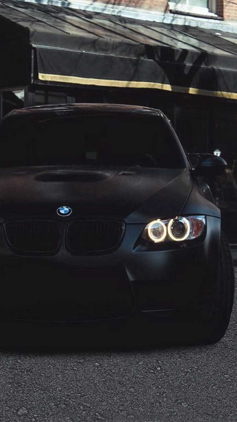Matte black BMW M3 with illuminated angel eyes headlights parked on asphalt against a blurred brick building.