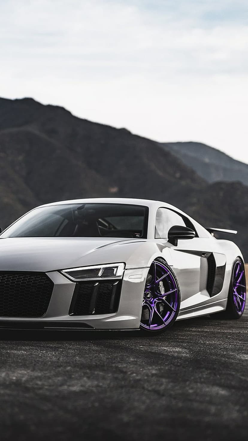 White Audi R8 supercar with custom purple rims parked on a mountain road against a dramatic rocky backdrop.