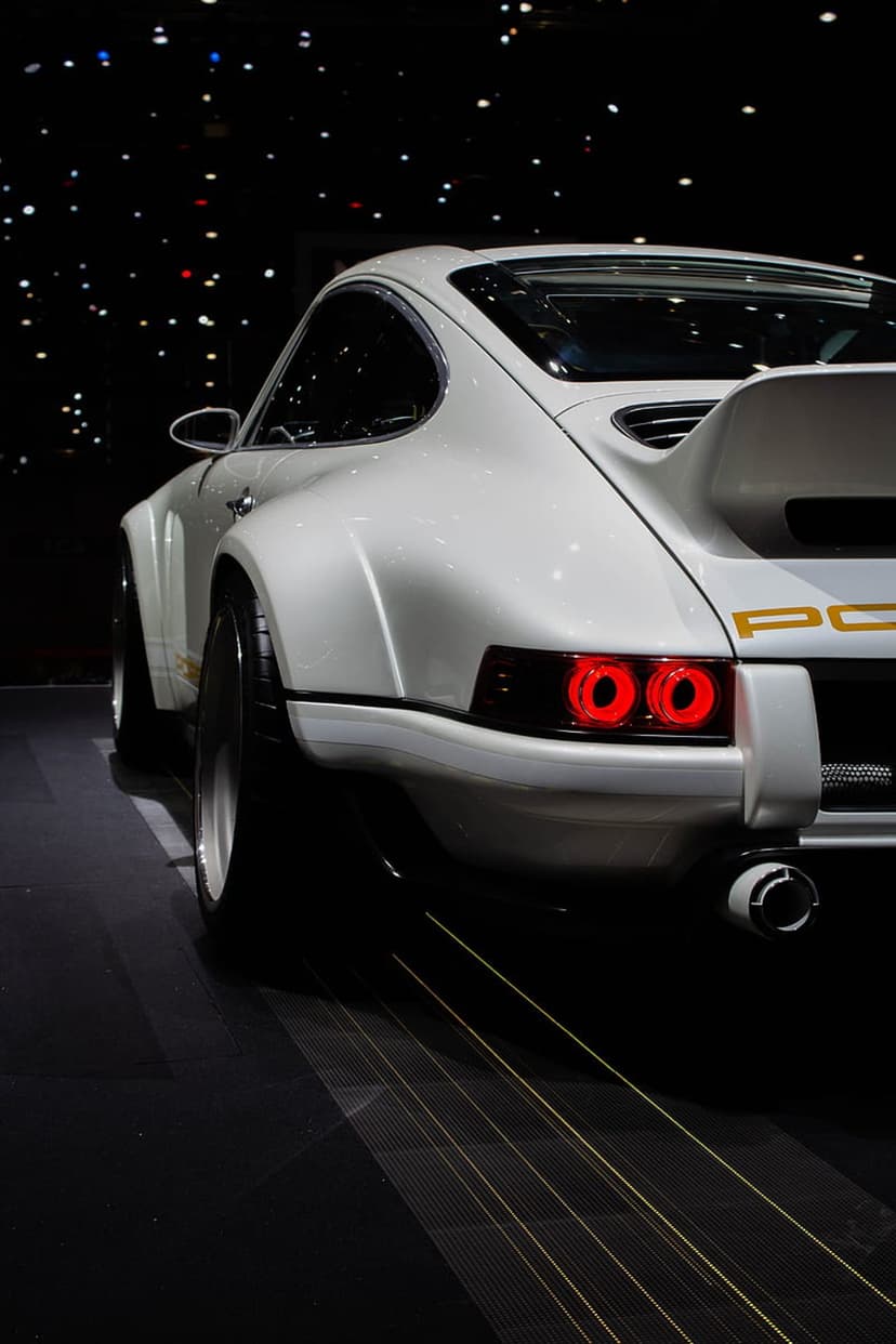 Porsche 911 rear view with white paint, red circular lights, and gold lettering against a dark night background.